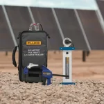 Field setup with solar panels in the background; a Fluke SOLMETRIC I-V curve tracer on rocky ground, with its blue handheld probe nearby on the ground.